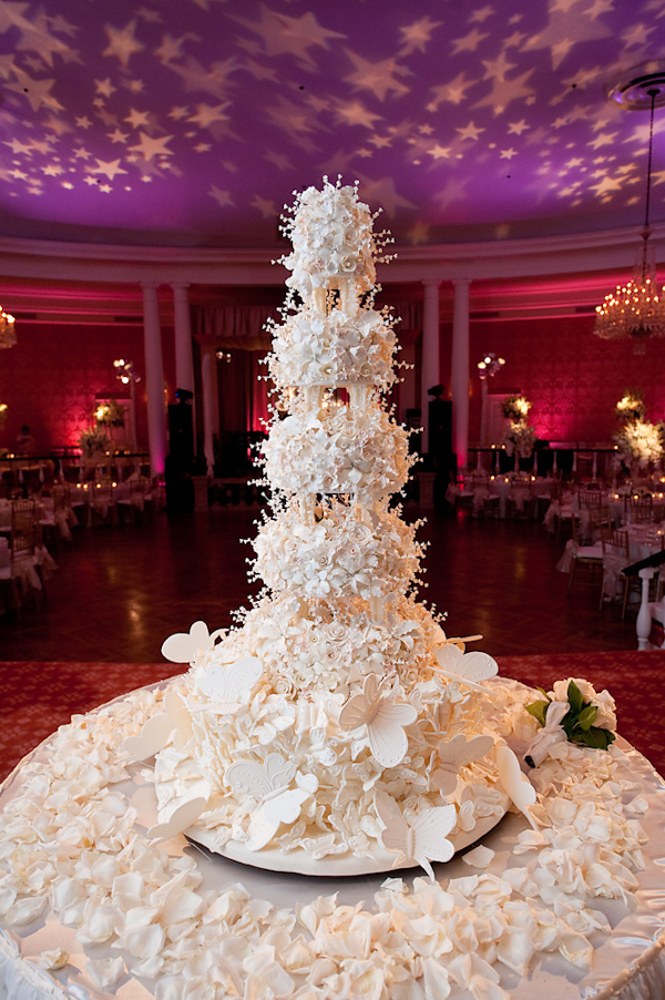 photo of a beautiful five tier ivory wedding cake with floral decorations surrounded by ivory flower petals on a round cake table at the reception with pink and purple lighting in the background with stars on the ceiling - photo by Houston based wedding photographer Adam Nyholt 